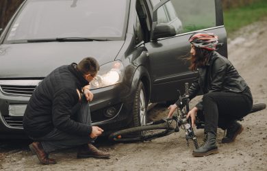 Man and woman inspecting car and bicycle after roadside accident, both crouched down assessing damage