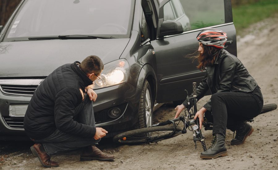 Man and woman inspecting car and bicycle after roadside accident, both crouched down assessing damage