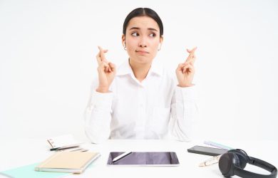 Person in white shirt sits at a desk with crossed fingers, surrounded by a tablet, notebooks, and headphones