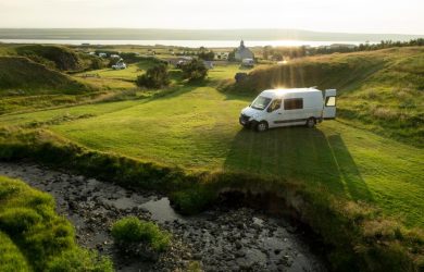 A camper van parked on a grassy hillside, surrounded by lush greenery and a clear blue sky