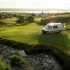 A camper van parked on a grassy hillside, surrounded by lush greenery and a clear blue sky