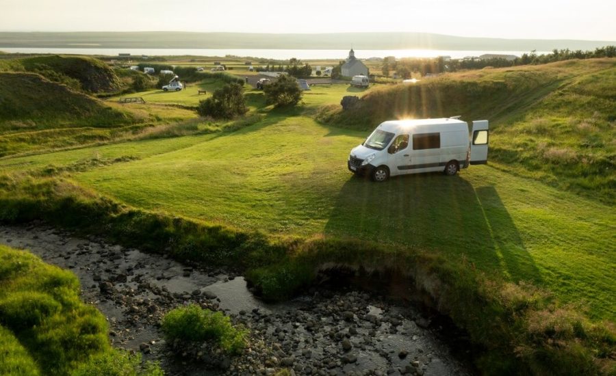 A camper van parked on a grassy hillside, surrounded by lush greenery and a clear blue sky