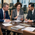 Three business professionals discussing reports at a meeting table in a modern office setting.